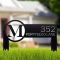 Metal address sign featuring an “M” logo displayed on grass in front of a house, highlighting durable craftsmanship and decorative outdoor appeal.