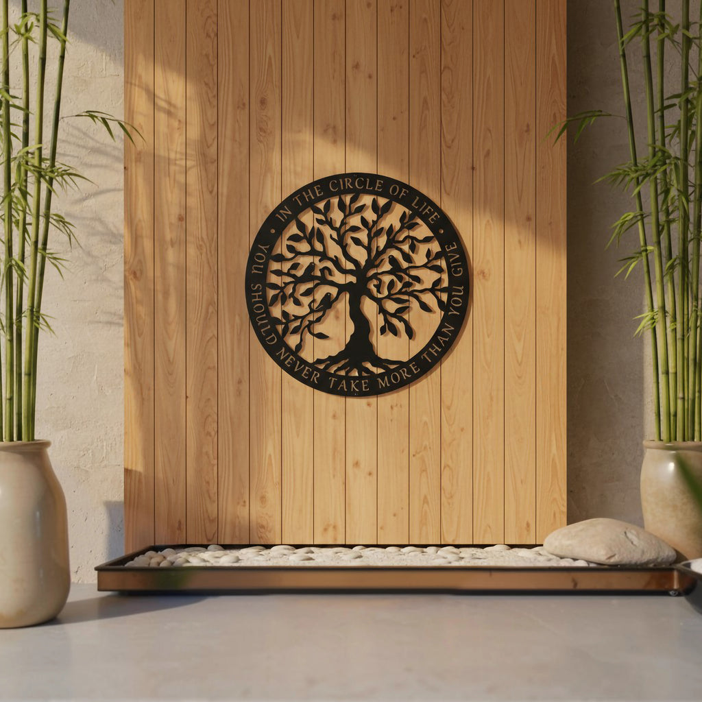 A satin black circular metal sign displaying a detailed Tree of Life and an embedded life quote is centered on a vertical light-wood plank feature wall. Sunlight casts shadows across the display, which is framed by tall potted bamboo stalks and a tray of white river stones.