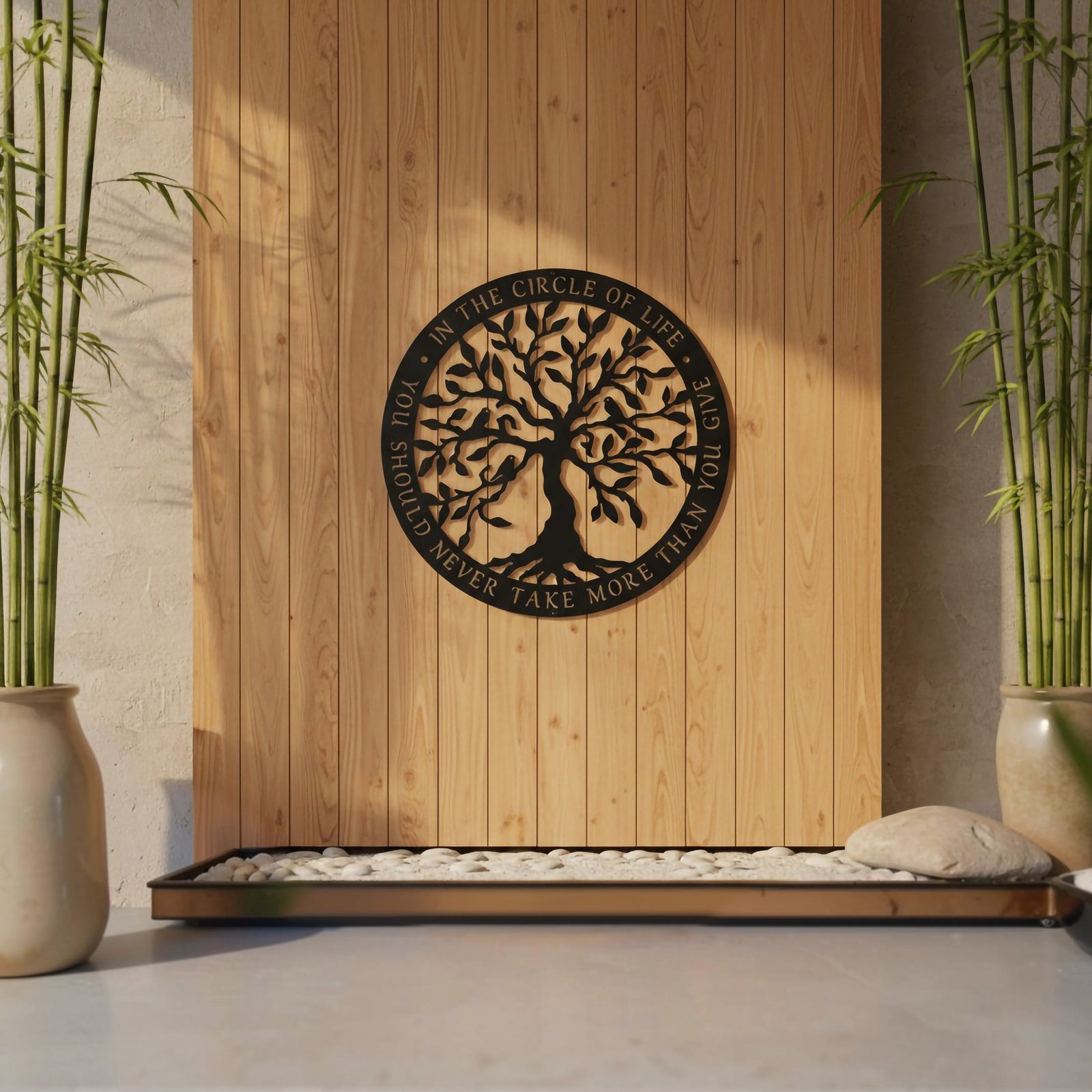A satin black circular metal sign displaying a detailed Tree of Life and an embedded life quote is centered on a vertical light-wood plank feature wall. Sunlight casts shadows across the display, which is framed by tall potted bamboo stalks and a tray of white river stones.