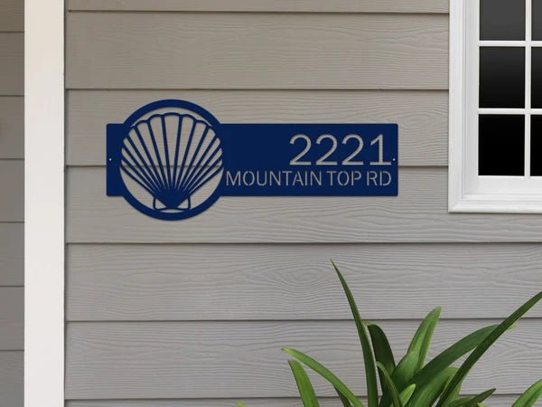 Seashell house number sign on gray siding wall, personalized metal address plaque, coastal front door décor, outdoor nautical home sign.