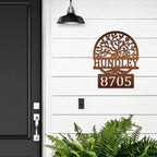 Decorative metal house number sign with tree design on white wall next to black door, custom outdoor address plaque, modern home exterior décor