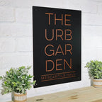 Black metal sign displaying “THE URB GARDEN MERCANTILE HALL” mounted on a white brick wall, highlighting bold lettering and a clean, professional design.