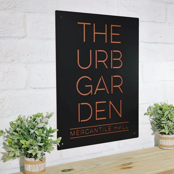 Black metal sign displaying “THE URB GARDEN MERCANTILE HALL” mounted on a white brick wall, highlighting bold lettering and a clean, professional design.