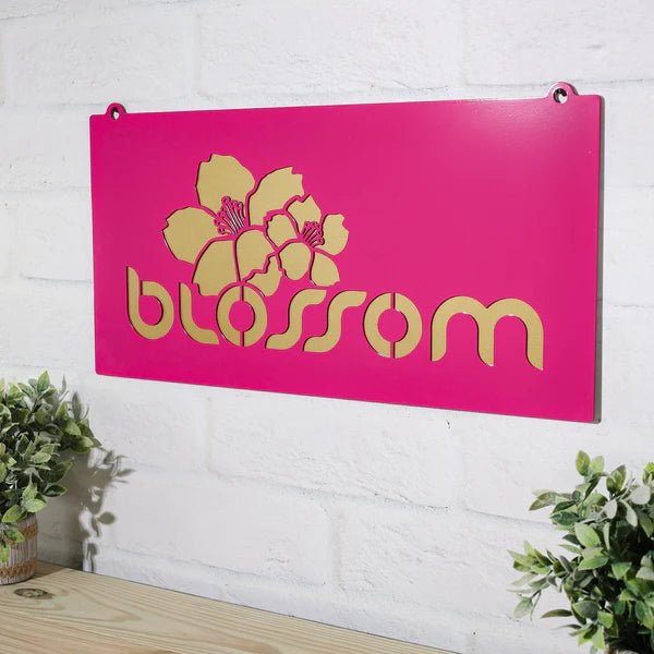 Pink metal sign featuring a yellow floral design and the text “blossom” mounted on a white brick wall, showcasing vibrant colors and decorative, eye-catching design.