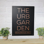 Black metal sign featuring the text “THE URB GARDEN MERCANTILE HALL” mounted on a white brick wall, showcasing bold lettering and a sleek, professional design.