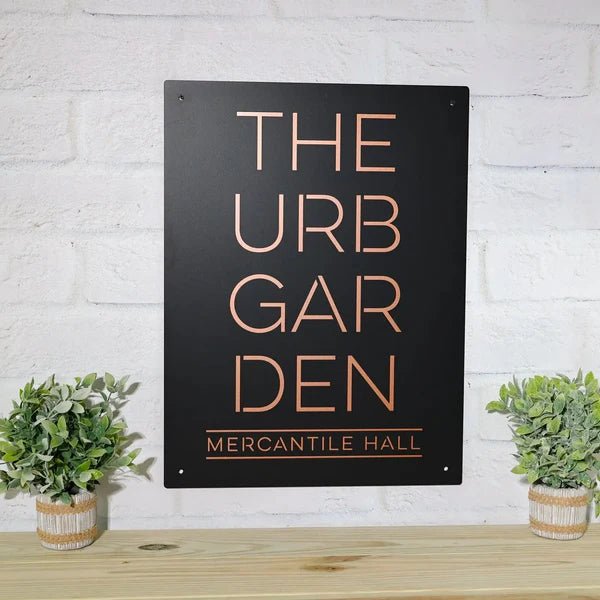 Black metal sign featuring the text “THE URB GARDEN MERCANTILE HALL” mounted on a white brick wall, showcasing bold lettering and a sleek, professional design.