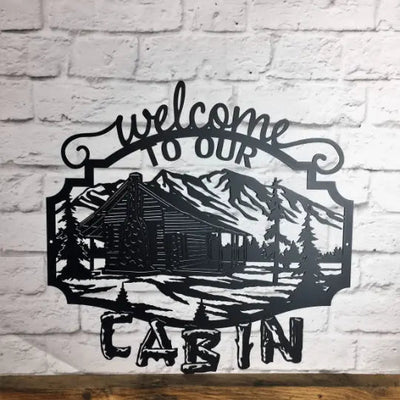 Black rustic “Welcome to Our Cabin” metal wall sign with cabin illustration on white brick wall, custom powder coated farmhouse décor, entry welcome sign design