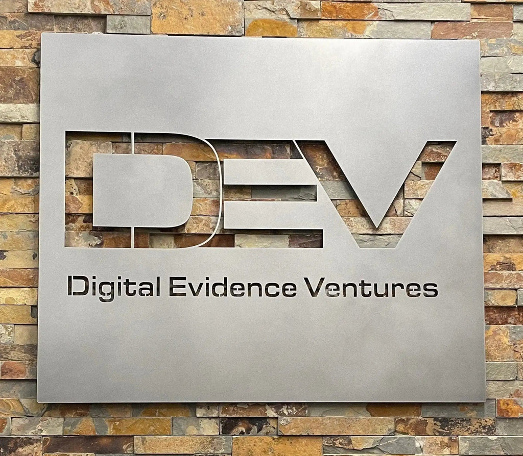 HouseSensationsArt custom metal sign with ‘Digital Evidence Ventures’ logo mounted on a stone wall using Weld Mount System strong adhesive fasteners, matte black powder-coated finish, durable outdoor-ready business signage, modern metal wall décor.