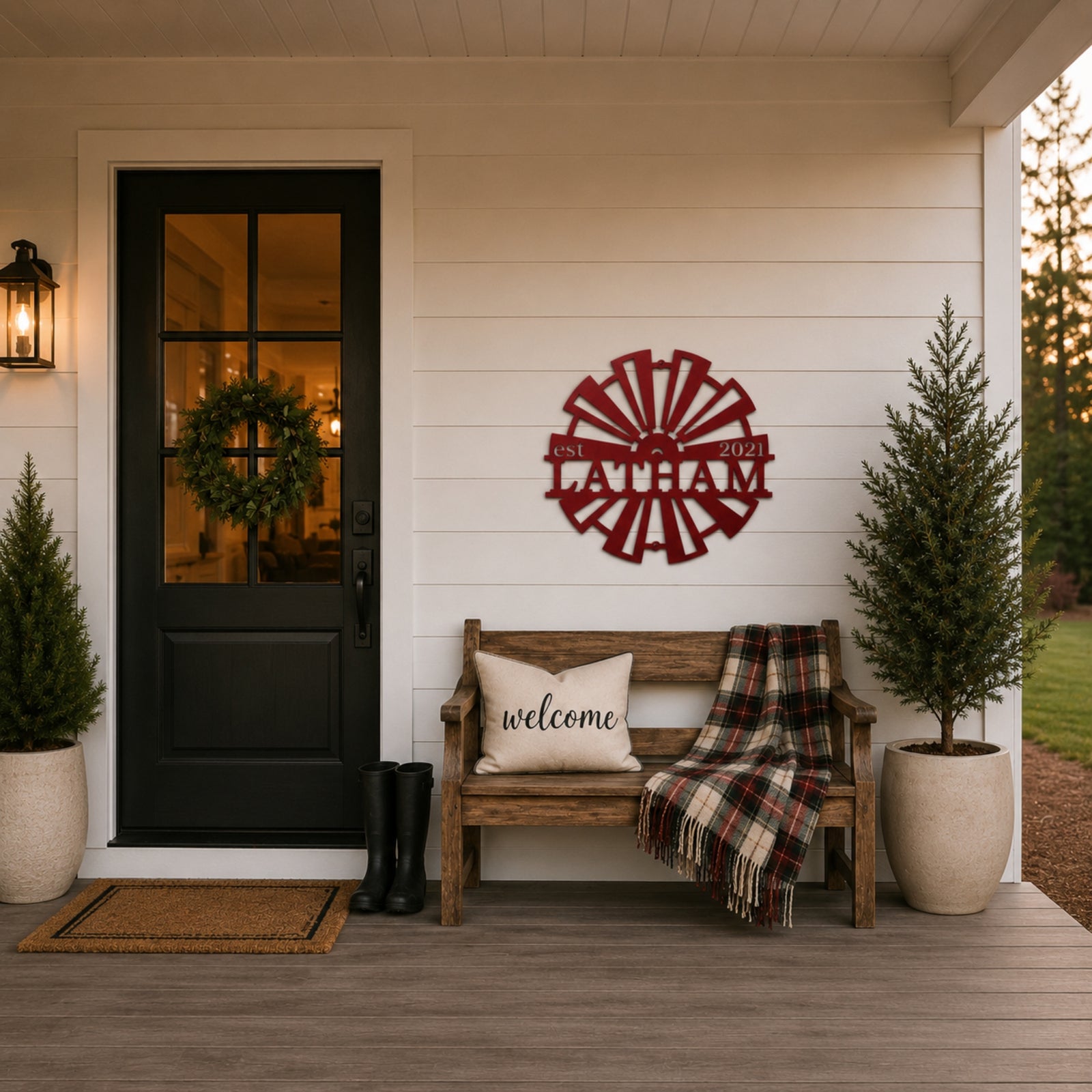 Red customized windmill metal wall art with "Latham est 2021" mounted on a white farmhouse porch wall above a wooden bench with a "welcome" pillow and plaid blanket.