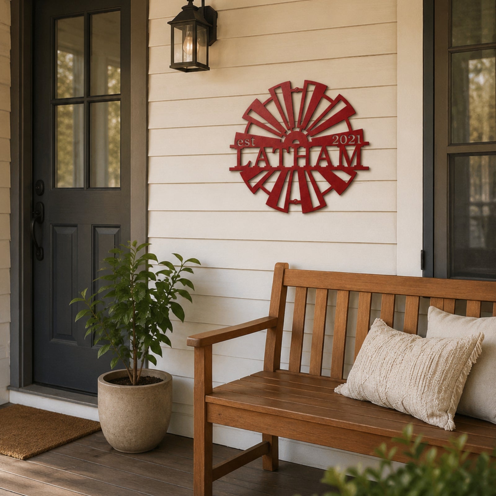 Custom red metal windmill wall art with "Latham est 2021" nameplate, displayed on a cream farmhouse exterior wall above a wooden garden bench with decorative pillows.