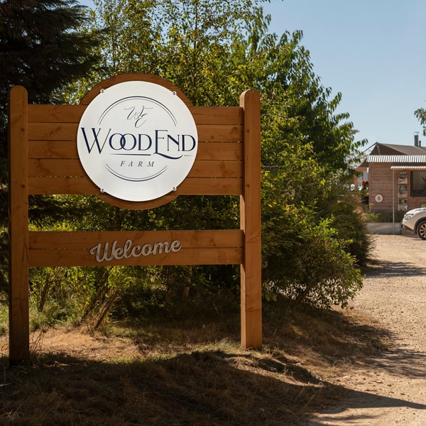 Wooden sign with 'WoodEnd Farm' logo and 'Welcome' text, surrounded by greenery.