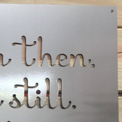 Close-up of a metal sign with cut-out letters spelling “then. still.” displayed on a wooden surface, highlighting detailed craftsmanship and modern decorative design.