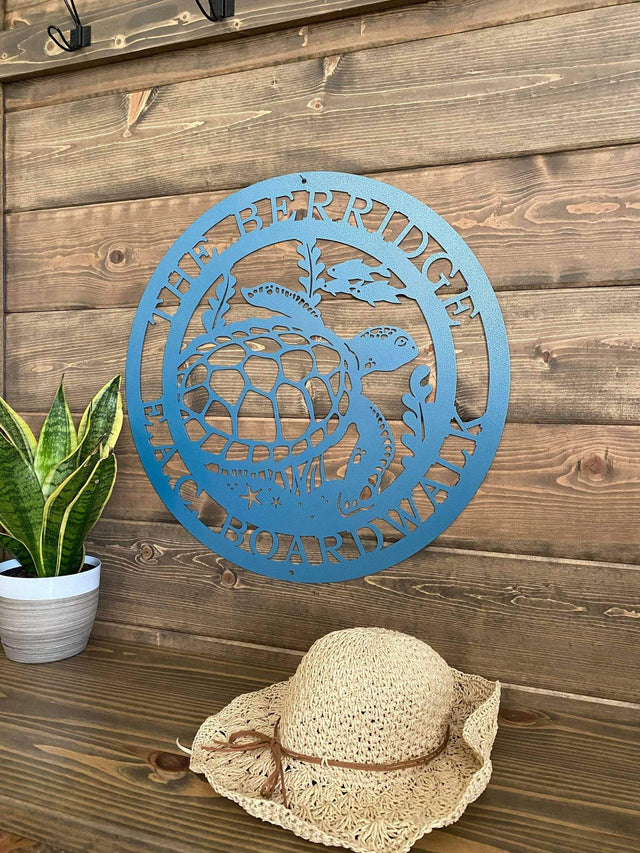 Angled view of textured blue custom laser cut metal sign showing turtle, seaweed, and fish, with “THE BERRIDGE E.A.C BOARDWALK” text, mounted on wood wall.